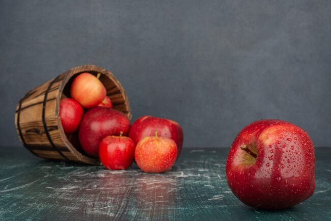 Red apples falling out of bucket on marble table Apples in a wooden bucket with apple on the foreground illustrate blog "Apples: What Vitamins and Minerals Do They Contain?"